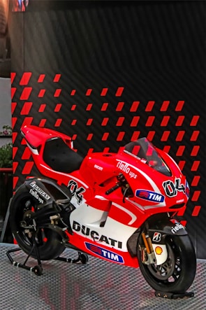 A red and white Ducati racing motorcycle is displayed on a metallic platform. It is adorned with various sponsor logos, including TIM, Shell, and Bridgestone, and features the number 04. The background consists of diagonal red and black patterned panels.