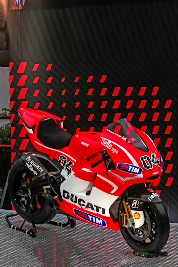 A red and white Ducati racing motorcycle is displayed on a metallic platform. It is adorned with various sponsor logos, including TIM, Shell, and Bridgestone, and features the number 04. The background consists of diagonal red and black patterned panels.