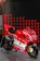 A red and white Ducati racing motorcycle is displayed on a metallic platform. It is adorned with various sponsor logos, including TIM, Shell, and Bridgestone, and features the number 04. The background consists of diagonal red and black patterned panels.