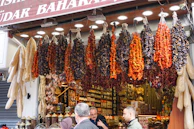 A bustling market stall is displayed with an array of hanging dried vegetables, including red peppers and dried eggplants. Below the hanging display, jars of spices are neatly arranged. Shoppers are engaging in conversation, creating a lively and vibrant scene.