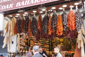 A bustling market stall is displayed with an array of hanging dried vegetables, including red peppers and dried eggplants. Below the hanging display, jars of spices are neatly arranged. Shoppers are engaging in conversation, creating a lively and vibrant scene.