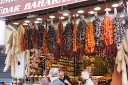 A bustling market stall is displayed with an array of hanging dried vegetables, including red peppers and dried eggplants. Below the hanging display, jars of spices are neatly arranged. Shoppers are engaging in conversation, creating a lively and vibrant scene.