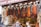 A bustling market stall is displayed with an array of hanging dried vegetables, including red peppers and dried eggplants. Below the hanging display, jars of spices are neatly arranged. Shoppers are engaging in conversation, creating a lively and vibrant scene.