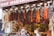 A bustling market stall is displayed with an array of hanging dried vegetables, including red peppers and dried eggplants. Below the hanging display, jars of spices are neatly arranged. Shoppers are engaging in conversation, creating a lively and vibrant scene.
