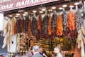 A bustling market stall is displayed with an array of hanging dried vegetables, including red peppers and dried eggplants. Below the hanging display, jars of spices are neatly arranged. Shoppers are engaging in conversation, creating a lively and vibrant scene.
