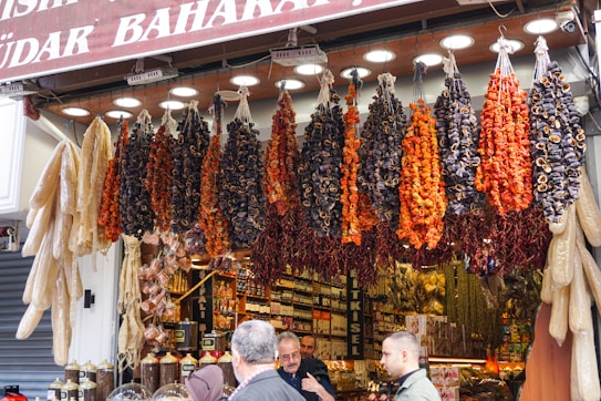 A bustling market stall is displayed with an array of hanging dried vegetables, including red peppers and dried eggplants. Below the hanging display, jars of spices are neatly arranged. Shoppers are engaging in conversation, creating a lively and vibrant scene.