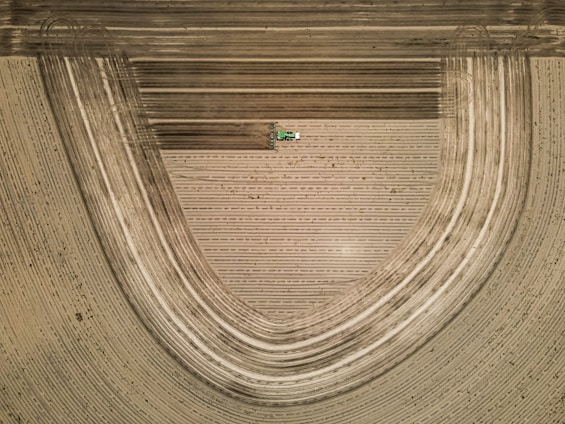 Aerial view of a tractor working on agricultural fields, creating intricate and curved patterns in the soil. The landscape is composed of parallel lines with a distinct curvature, indicating efficient land preparation and cultivation.