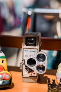 A vintage Bell & Howell camera with a distinctive three-lens design is prominently displayed. It is surrounded by colorful, artistic objects, including a toy car and a painted figure, placed on a wooden surface.