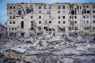 A severely damaged multi-story building with multiple broken windows and structural damage. Surrounding the building are piles of rubble and debris, including broken concrete and exposed metal. The scene indicates a recent event of destruction.