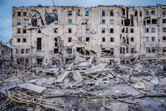 A severely damaged multi-story building with multiple broken windows and structural damage. Surrounding the building are piles of rubble and debris, including broken concrete and exposed metal. The scene indicates a recent event of destruction.