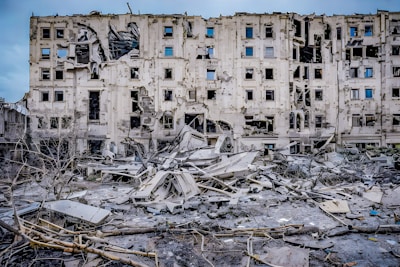 A severely damaged multi-story building with multiple broken windows and structural damage. Surrounding the building are piles of rubble and debris, including broken concrete and exposed metal. The scene indicates a recent event of destruction.