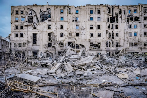 A severely damaged multi-story building with multiple broken windows and structural damage. Surrounding the building are piles of rubble and debris, including broken concrete and exposed metal. The scene indicates a recent event of destruction.
