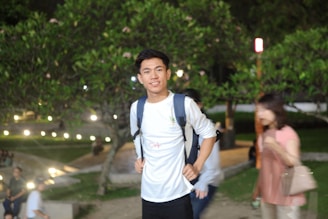 A young person with a backpack is standing outdoors, smiling at the camera. The background features greenery and blurred lights, suggesting a park or garden setting in the evening. Other individuals are visible but out of focus, indicating motion or camera lens effects.