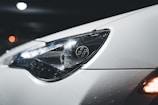 A close-up view of a car's headlight, showcasing its sleek design. The car surface appears white and is covered in small water droplets, suggesting wet conditions or recent washing. The headlight features a distinct logo with stylized numbers, while reflections play across the glossy surface. In the background, there are blurred bokeh lights, giving the image a night-time or low-light setting.