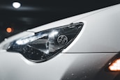 A close-up view of a car's headlight, showcasing its sleek design. The car surface appears white and is covered in small water droplets, suggesting wet conditions or recent washing. The headlight features a distinct logo with stylized numbers, while reflections play across the glossy surface. In the background, there are blurred bokeh lights, giving the image a night-time or low-light setting.
