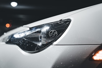 A close-up view of a car's headlight, showcasing its sleek design. The car surface appears white and is covered in small water droplets, suggesting wet conditions or recent washing. The headlight features a distinct logo with stylized numbers, while reflections play across the glossy surface. In the background, there are blurred bokeh lights, giving the image a night-time or low-light setting.