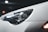 A close-up view of a car's headlight, showcasing its sleek design. The car surface appears white and is covered in small water droplets, suggesting wet conditions or recent washing. The headlight features a distinct logo with stylized numbers, while reflections play across the glossy surface. In the background, there are blurred bokeh lights, giving the image a night-time or low-light setting.