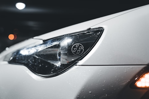 A close-up view of a car's headlight, showcasing its sleek design. The car surface appears white and is covered in small water droplets, suggesting wet conditions or recent washing. The headlight features a distinct logo with stylized numbers, while reflections play across the glossy surface. In the background, there are blurred bokeh lights, giving the image a night-time or low-light setting.