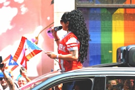 A person with long black hair and a red football jersey is standing out of a car roof holding a microphone. They are surrounded by people and waving a small Puerto Rican flag. In the background, there's a rainbow flag displayed on a building.