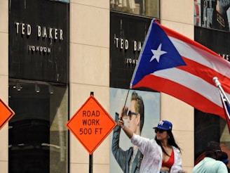 A woman wearing a red top and blue hat is waving a Puerto Rican flag in front of a Ted Baker London store. Two road work signs, reading 'ROAD WORK 500 FT', are visible nearby, suggesting construction in the area.
