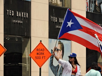 A woman wearing a red top and blue hat is waving a Puerto Rican flag in front of a Ted Baker London store. Two road work signs, reading 'ROAD WORK 500 FT', are visible nearby, suggesting construction in the area.