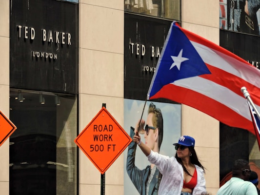 A woman wearing a red top and blue hat is waving a Puerto Rican flag in front of a Ted Baker London store. Two road work signs, reading 'ROAD WORK 500 FT', are visible nearby, suggesting construction in the area.