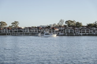 A serene waterfront scene featuring a large, white motorboat navigating on calm blue waters. In the background, there is a row of modern, multi-story houses with red roofs, nestled among tall trees. The sky is clear, suggesting a peaceful day.