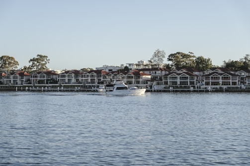 A serene waterfront scene featuring a large, white motorboat navigating on calm blue waters. In the background, there is a row of modern, multi-story houses with red roofs, nestled among tall trees. The sky is clear, suggesting a peaceful day.