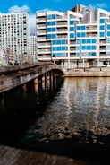 A modern apartment building with a reflective glass facade is located alongside a body of water. A wooden bridge stretches over the water, leading towards the building. Blue skies with scattered clouds are visible, and sunlight enhances the reflections in the windows and on the water’s surface.