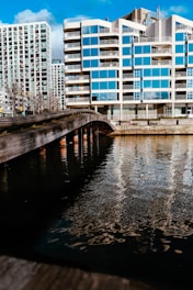 A modern apartment building with a reflective glass facade is located alongside a body of water. A wooden bridge stretches over the water, leading towards the building. Blue skies with scattered clouds are visible, and sunlight enhances the reflections in the windows and on the water’s surface.