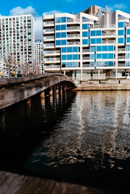 A modern apartment building with a reflective glass facade is located alongside a body of water. A wooden bridge stretches over the water, leading towards the building. Blue skies with scattered clouds are visible, and sunlight enhances the reflections in the windows and on the water’s surface.