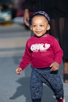 A young child is happily walking on a paved surface. The child is wearing a red sweatshirt with the words 'ONLY FOR YOU' printed on it and patterned pants. A hat with a blue and white anchor pattern adorns their head. In the background, an adult's leg is partially visible, and there is a blurred view of cars and a street.