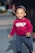 A young child is happily walking on a paved surface. The child is wearing a red sweatshirt with the words 'ONLY FOR YOU' printed on it and patterned pants. A hat with a blue and white anchor pattern adorns their head. In the background, an adult's leg is partially visible, and there is a blurred view of cars and a street.