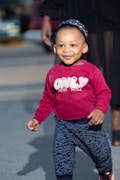A young child is happily walking on a paved surface. The child is wearing a red sweatshirt with the words 'ONLY FOR YOU' printed on it and patterned pants. A hat with a blue and white anchor pattern adorns their head. In the background, an adult's leg is partially visible, and there is a blurred view of cars and a street.