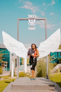 A woman stands on a platform in front of a decorative backdrop featuring large white wings, holding a graduation cap. The scene is set in a modern outdoor environment with green grass and blue-tinted glass buildings in the background. Above her, the structure displays '2022 GAU'.