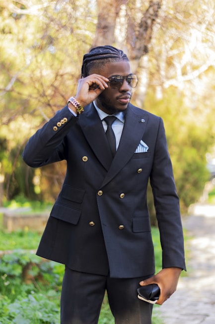 A fashionable man wearing a black double-breasted suit with gold buttons, paired with a white dress shirt and a black tie. He is adjusting his sunglasses with one hand while holding a phone in the other. The background consists of lush green foliage and a blurred pathway, giving a serene and stylish vibe.