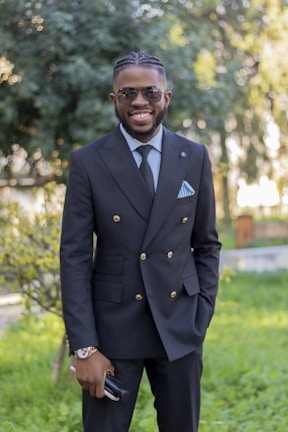 A man wearing a tailored blazer and leather watch, smiling while walking through a sunlit park.