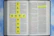 An open Bible with highlighted text in the book of John. Yellow tiles with blue letters are arranged in the shape of a cross spelling 'the good news.' The background is a soft blue sky pattern.