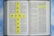 An open Bible with highlighted text in the book of John. Yellow tiles with blue letters are arranged in the shape of a cross spelling 'the good news.' The background is a soft blue sky pattern.