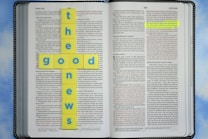 An open Bible with highlighted text in the book of John. Yellow tiles with blue letters are arranged in the shape of a cross spelling 'the good news.' The background is a soft blue sky pattern.