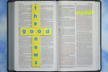 An open Bible with highlighted text in the book of John. Yellow tiles with blue letters are arranged in the shape of a cross spelling 'the good news.' The background is a soft blue sky pattern.