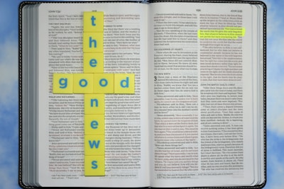 An open Bible with highlighted text in the book of John. Yellow tiles with blue letters are arranged in the shape of a cross spelling 'the good news.' The background is a soft blue sky pattern.
