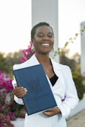 A person is smiling while holding a dark blue folder with an embossed logo and text. The individual is wearing a white outfit and has short hair. The background includes vibrant pink flowers and some greenery, suggesting an outdoor setting.