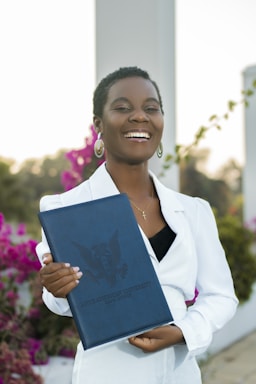 A person is smiling while holding a dark blue folder with an embossed logo and text. The individual is wearing a white outfit and has short hair. The background includes vibrant pink flowers and some greenery, suggesting an outdoor setting.