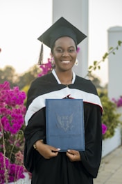 A graduate in a black cap and gown smiles while holding a diploma case. Vibrant pink flowers and greenery are in the background, along with architectural pillars.