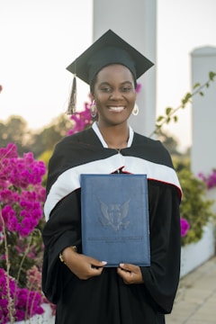 A graduate in a black cap and gown smiles while holding a diploma case. Vibrant pink flowers and greenery are in the background, along with architectural pillars.