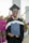 A graduate in a black cap and gown smiles while holding a diploma case. Vibrant pink flowers and greenery are in the background, along with architectural pillars.
