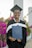 A graduate in a black cap and gown smiles while holding a diploma case. Vibrant pink flowers and greenery are in the background, along with architectural pillars.
