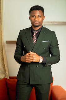 A person wearing a dark pinstripe suit with a plaid tie stands confidently. The background includes a light-colored wall and a partially visible orange sofa. The individual has short hair and is wearing a wristwatch.