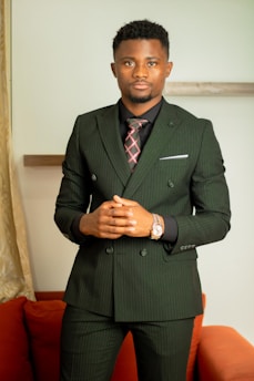 A person wearing a dark pinstripe suit with a plaid tie stands confidently. The background includes a light-colored wall and a partially visible orange sofa. The individual has short hair and is wearing a wristwatch.